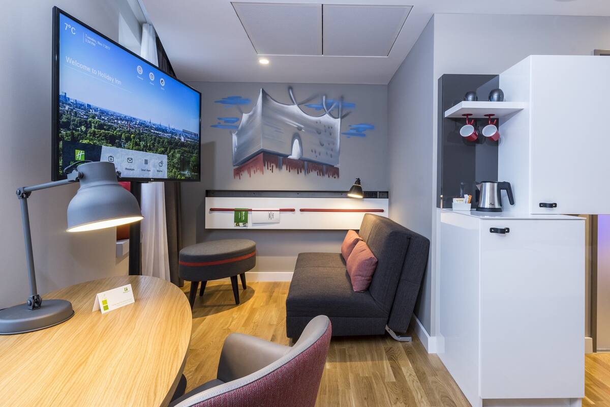 Une chambre d’hôtel moderne équipée d’un téléviseur à écran plat diffusant une vue sur la ville, d’un bureau en bois avec une lampe, d’un canapé gris agrémenté d’un coussin rose, d’une armoire blanche avec une bouilloire et d’une étagère murale où sont posées des tasses.