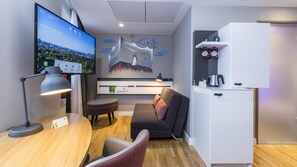 Ein modernes Hotelzimmer mit einem Flachbildfernseher, der eine Stadtansicht zeigt, einem Holztisch mit Lampe, einem grauen Sofa mit einem rosafarbenen Kissen, einem weißen Schrank mit Wasserkocher und einem Wandregal mit Tassen.