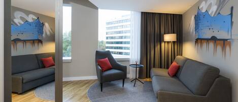 A modern hotel room with a grey sofa, a red pillow, a small round table, and a lamp. There is a large window with a view of a building and trees.