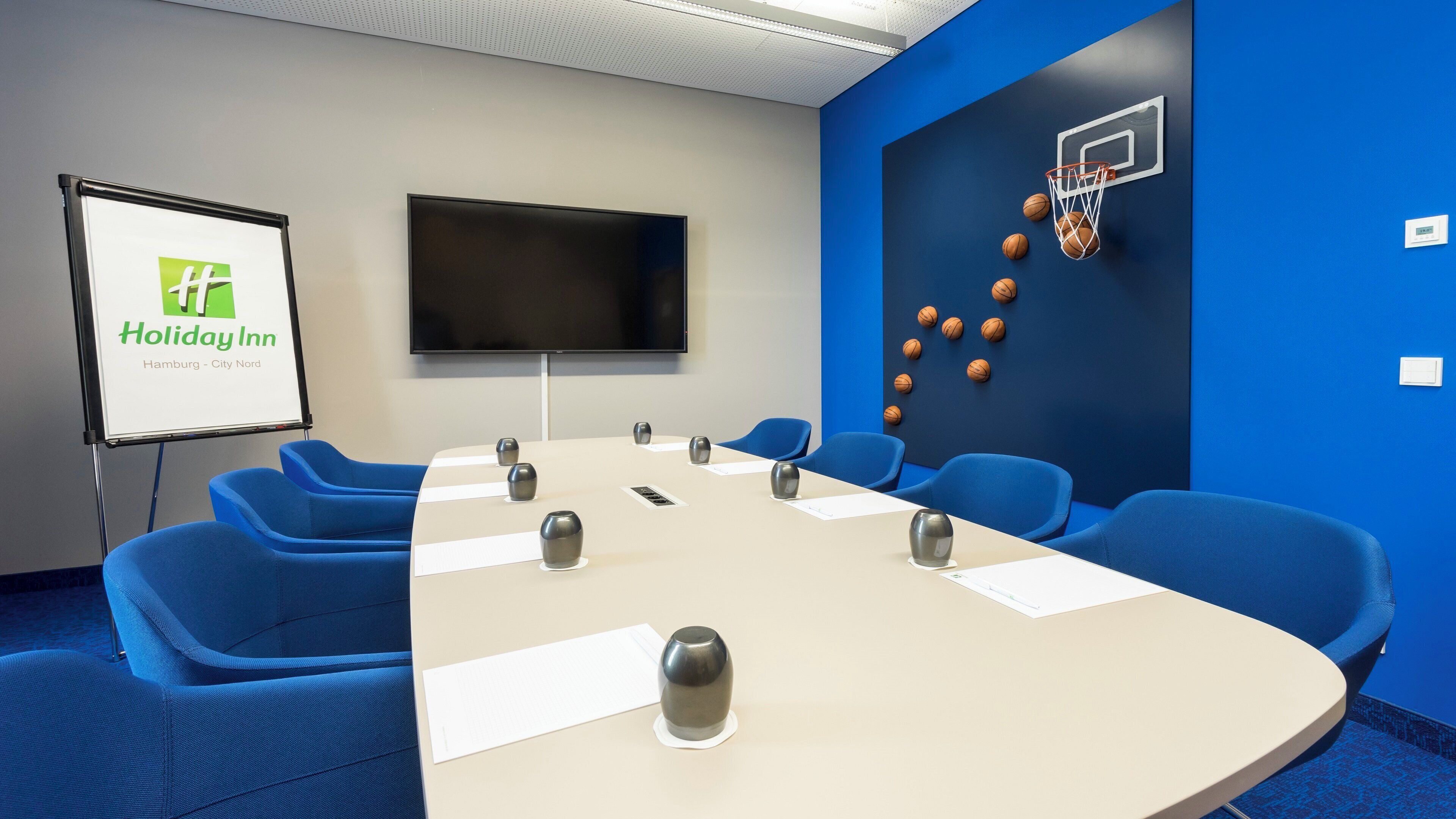 Un salón de conferencias con una larga mesa, sillas azules, una pizarra y un aro de baloncesto montado en la pared.