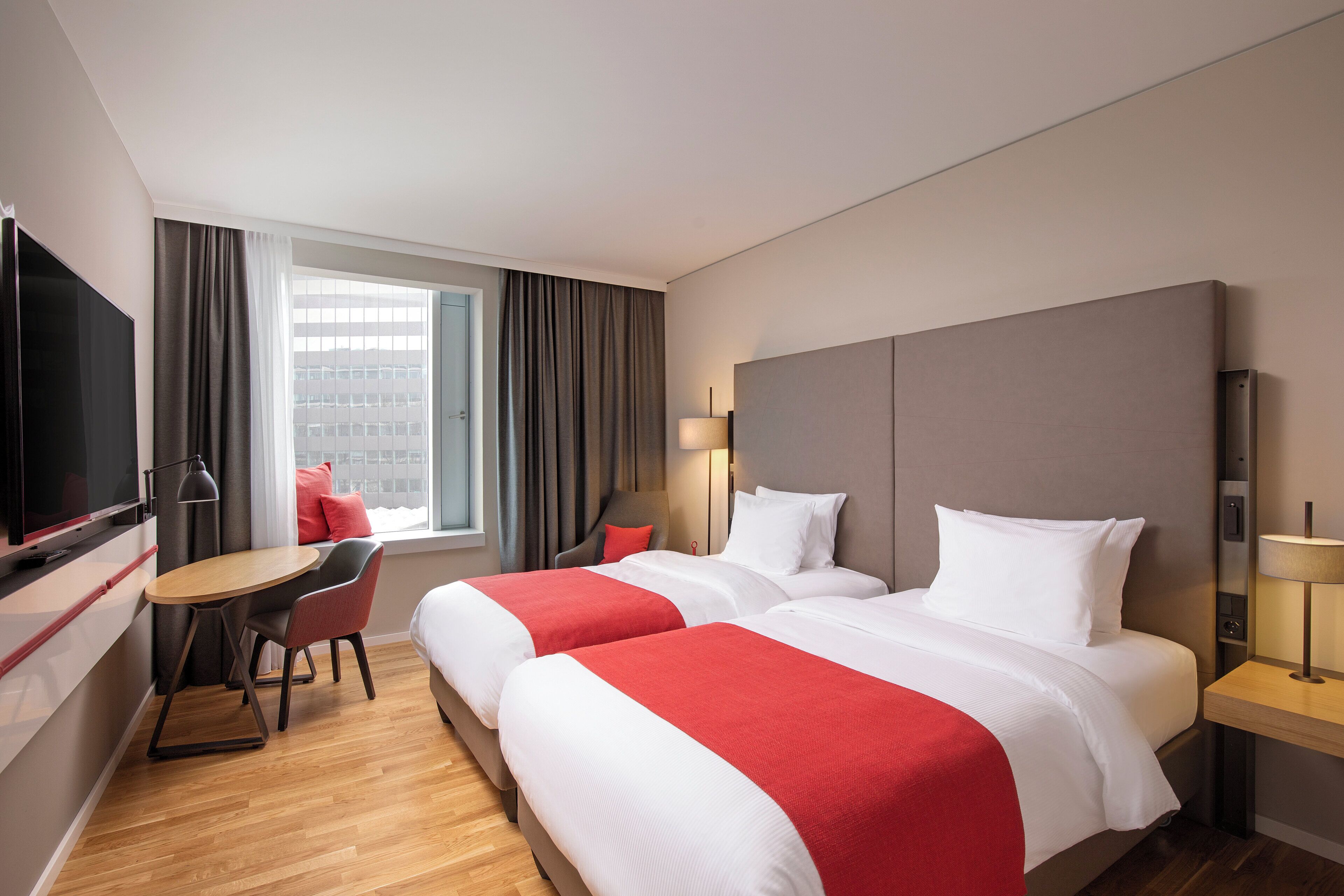 Une chambre d’hôtel avec deux lits, un bureau, une chaise, une télévision et une fenêtre avec des rideaux.