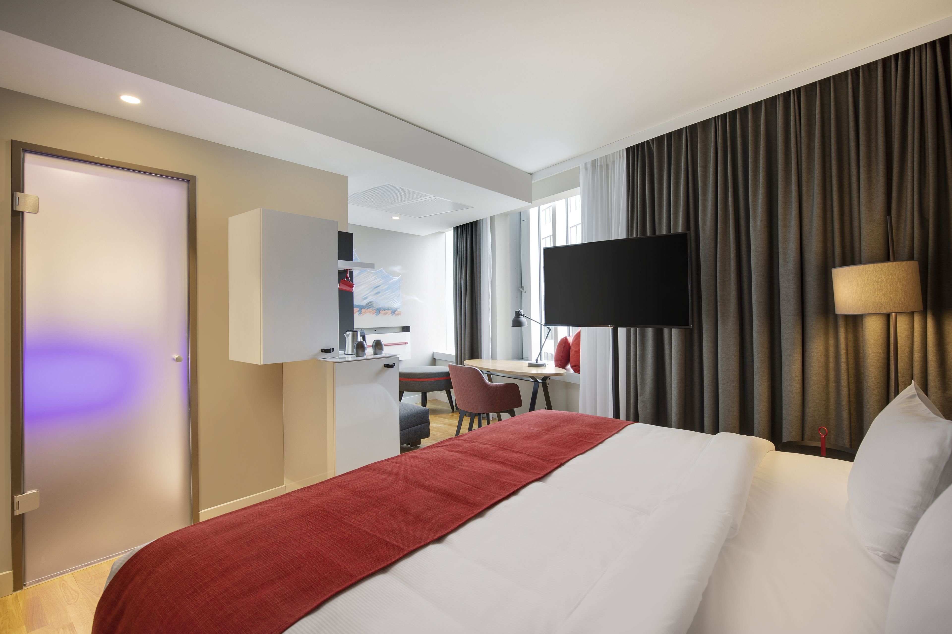 Une chambre d’hôtel avec un lit, un bureau, une chaise, une télévision et une fenêtre avec des rideaux.