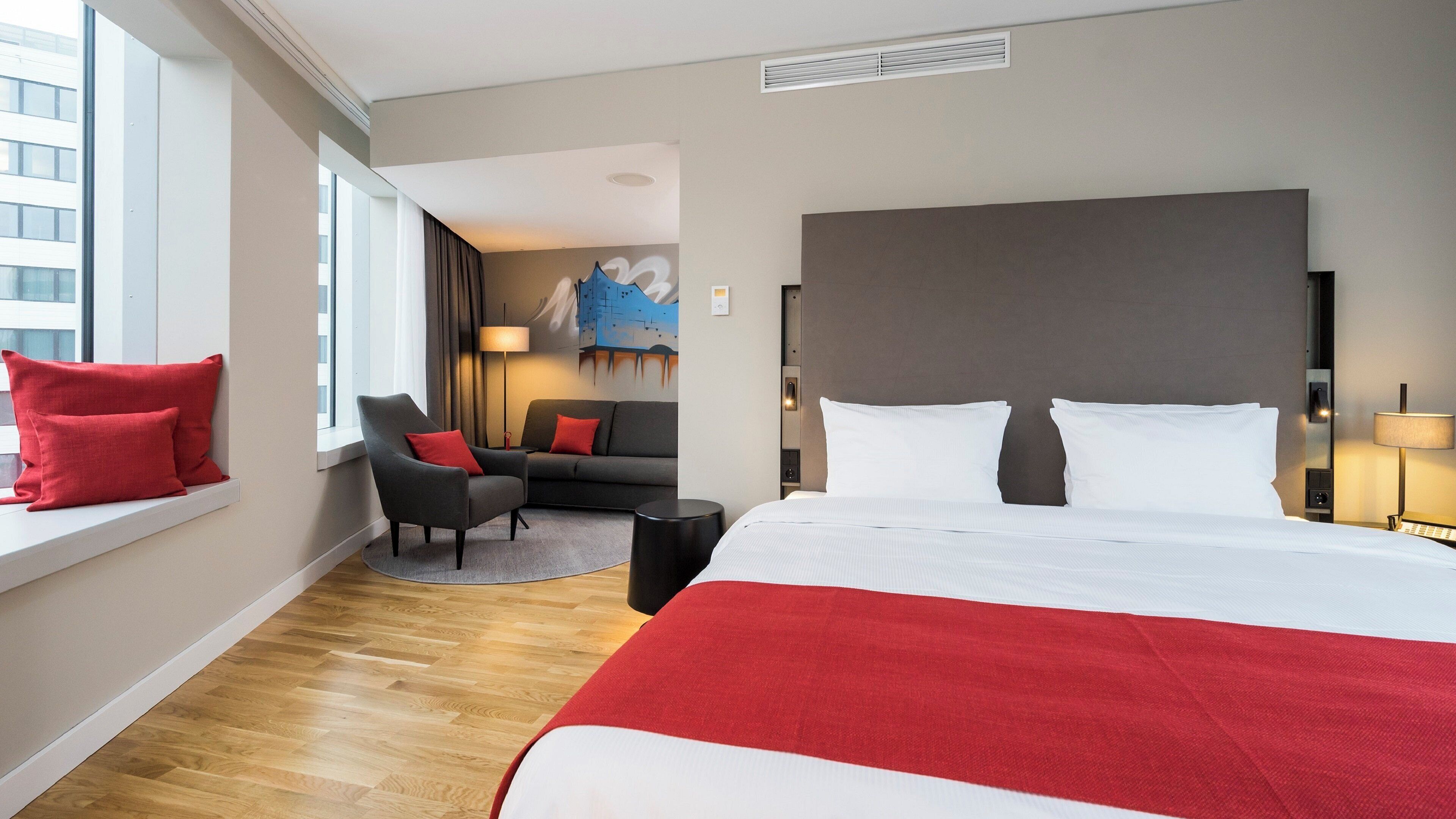 Habitación de hotel con una cama grande, un sofá, una silla y un ventanal con cortinas.