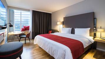 A hotel room with a large bed, a desk with a chair, a small table, and a view of the city through the window.