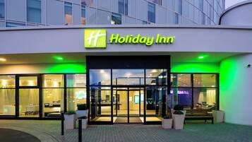 The exterior of a Holiday Inn hotel with a glass facade, a prominent sign, and a well-lit entrance.