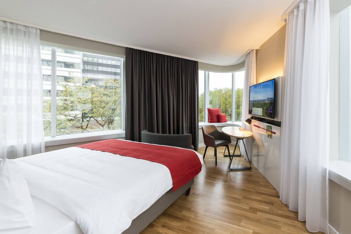 Une chambre d’hôtel avec un grand lit, un bureau avec une chaise, une télévision et une fenêtre avec des rideaux.