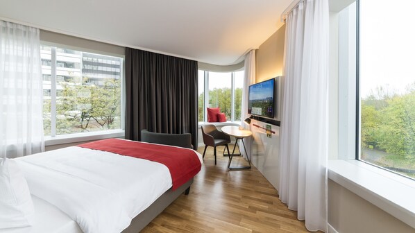 Ein Hotelzimmer mit einem großen Bett, einem Schreibtisch mit Stuhl, einem Fernseher und einem Fenster mit Vorhängen.