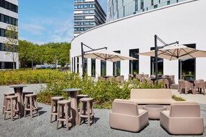 An outdoor seating area with tables and chairs, a sofa, and umbrellas, surrounded by greenery and modern buildings.