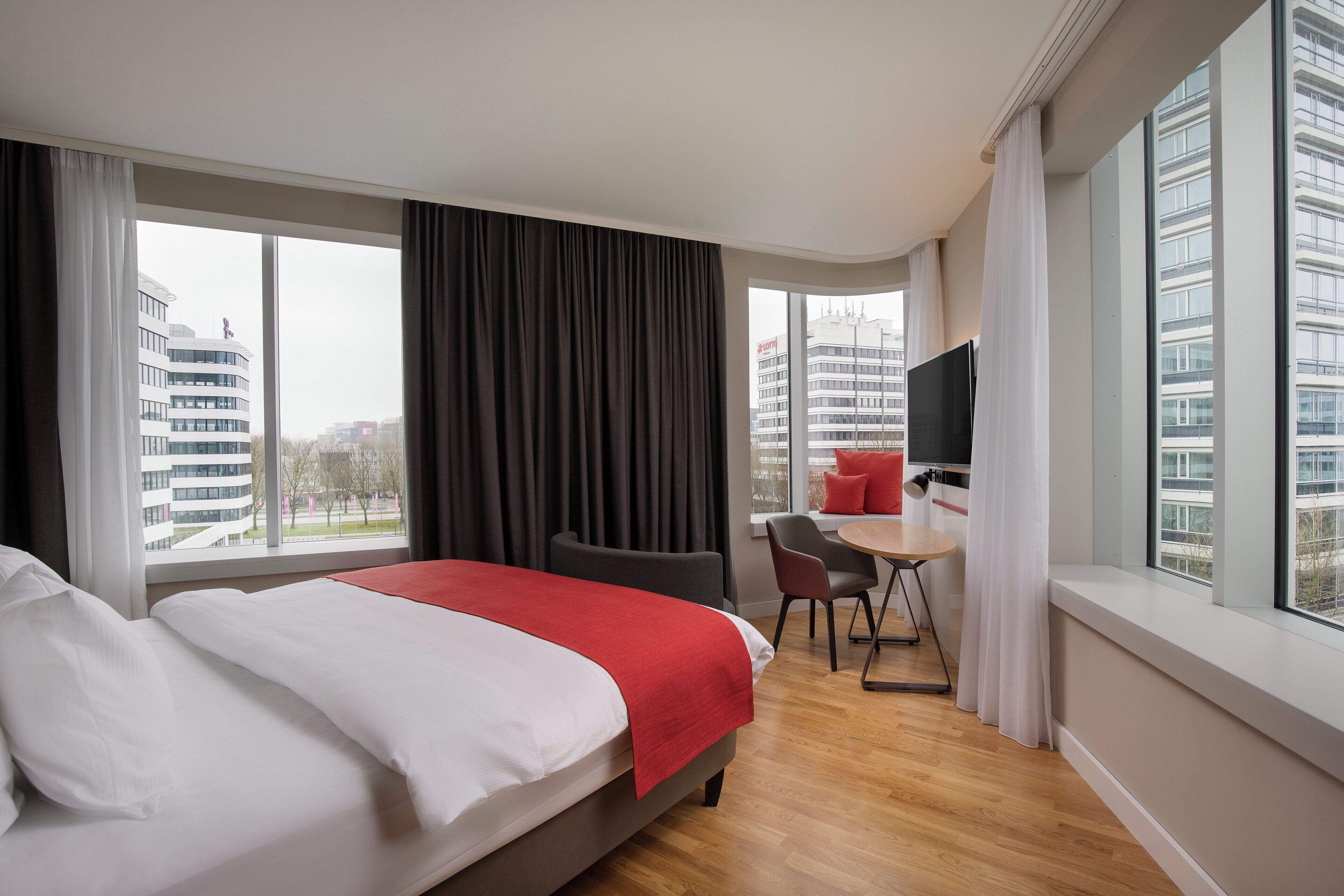 A hotel room with a bed, a desk, and a chair. There are large windows with curtains.