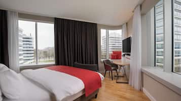 A hotel room with a bed, a desk, and a chair. There are large windows with curtains.