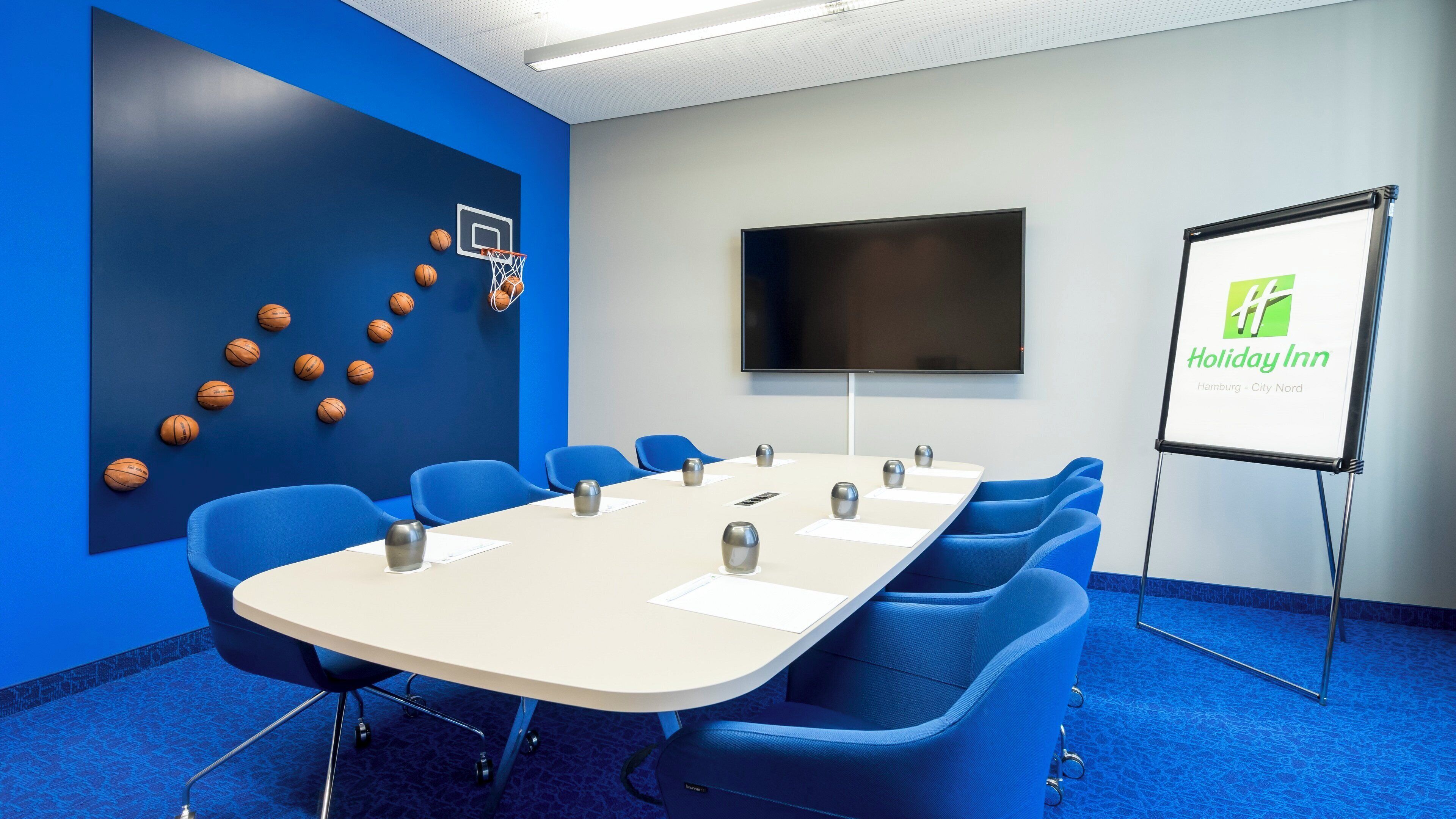 A conference room with a long table, blue chairs, a whiteboard, and a screen.