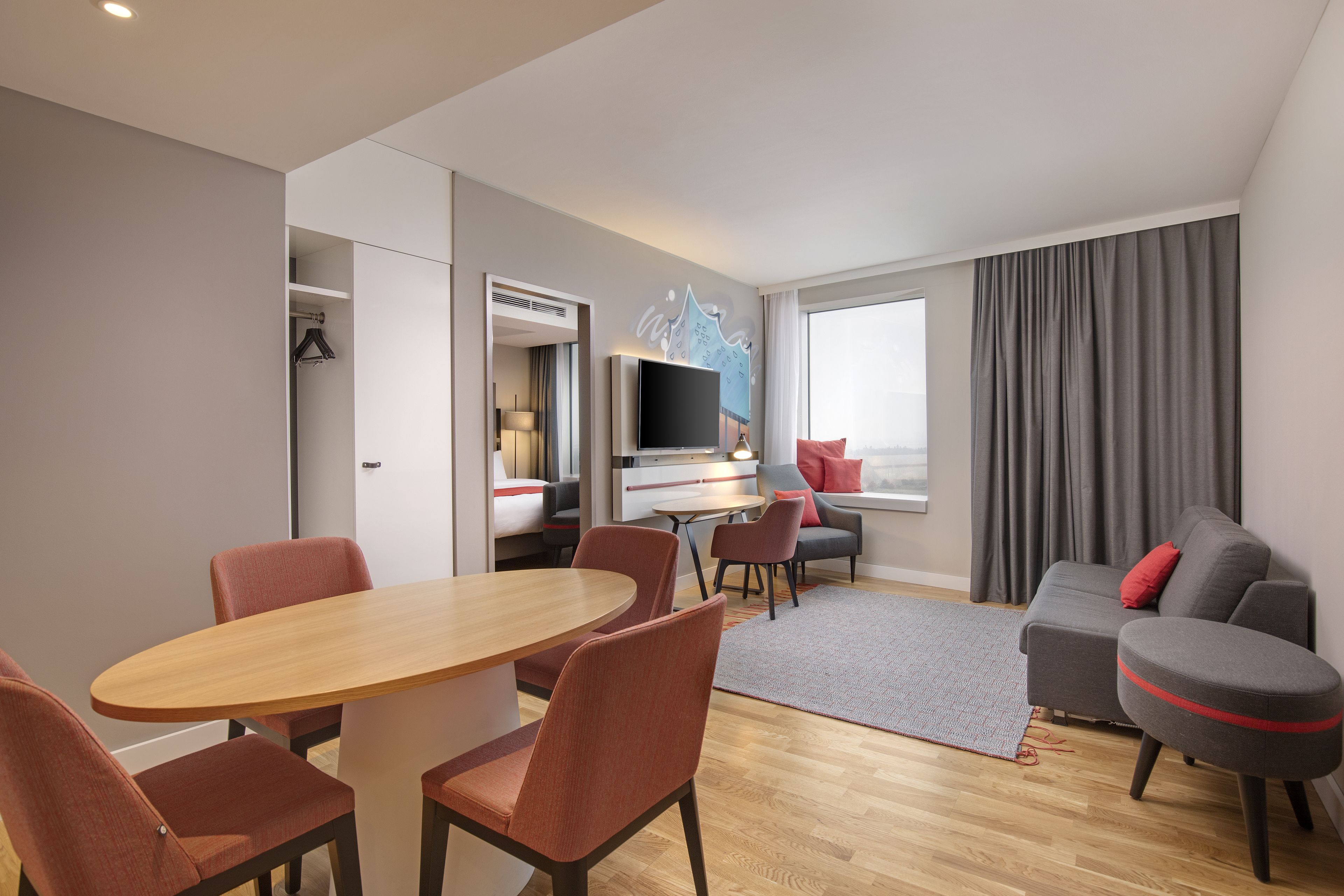 A modern hotel room with a dining area, a sofa, a TV, and a large window with curtains.