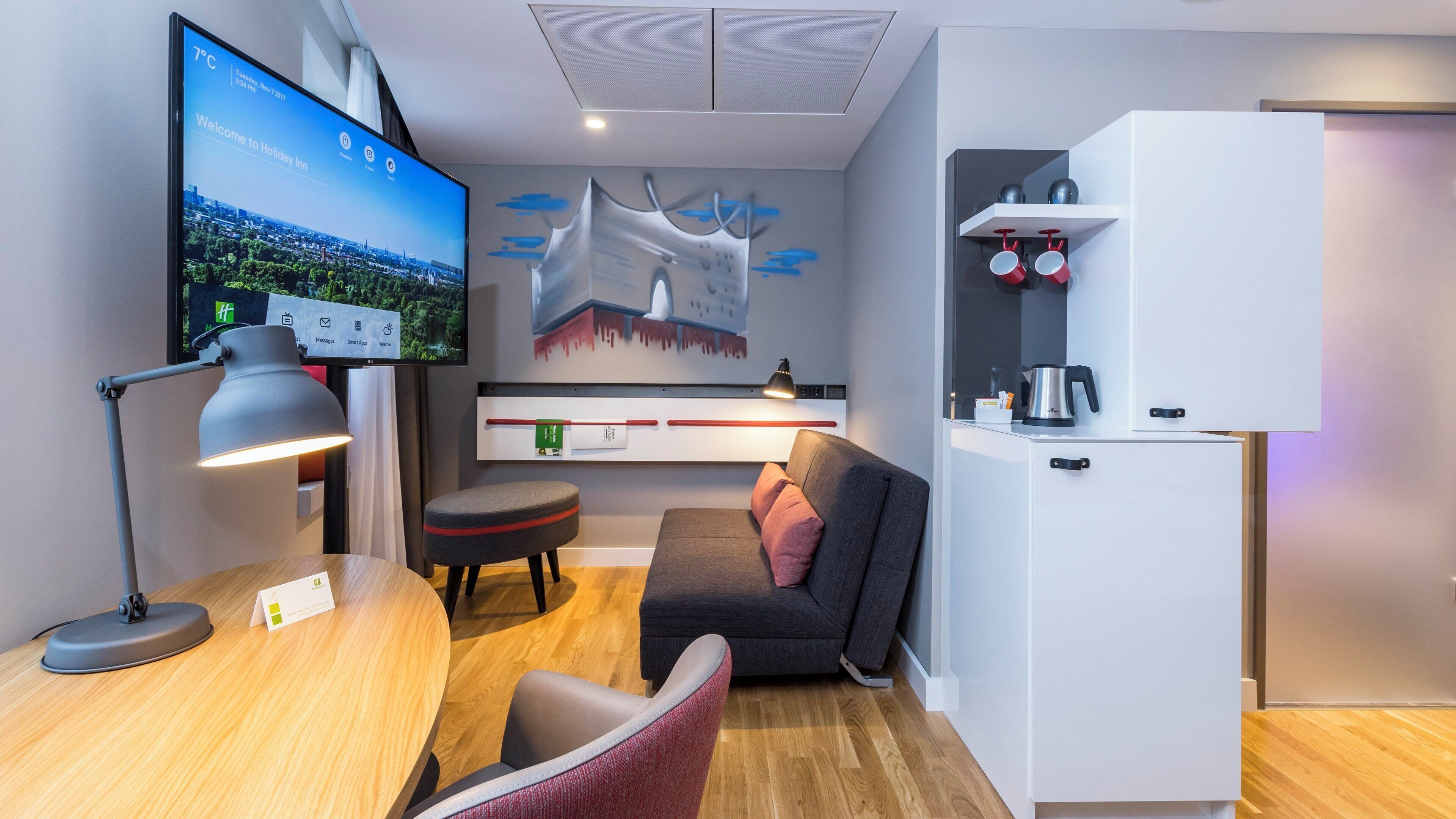 Een moderne hotelkamer met een flatscreen televisie, een bureau met een lamp, een sofa, een koffiezetapparaat en een koelkast.