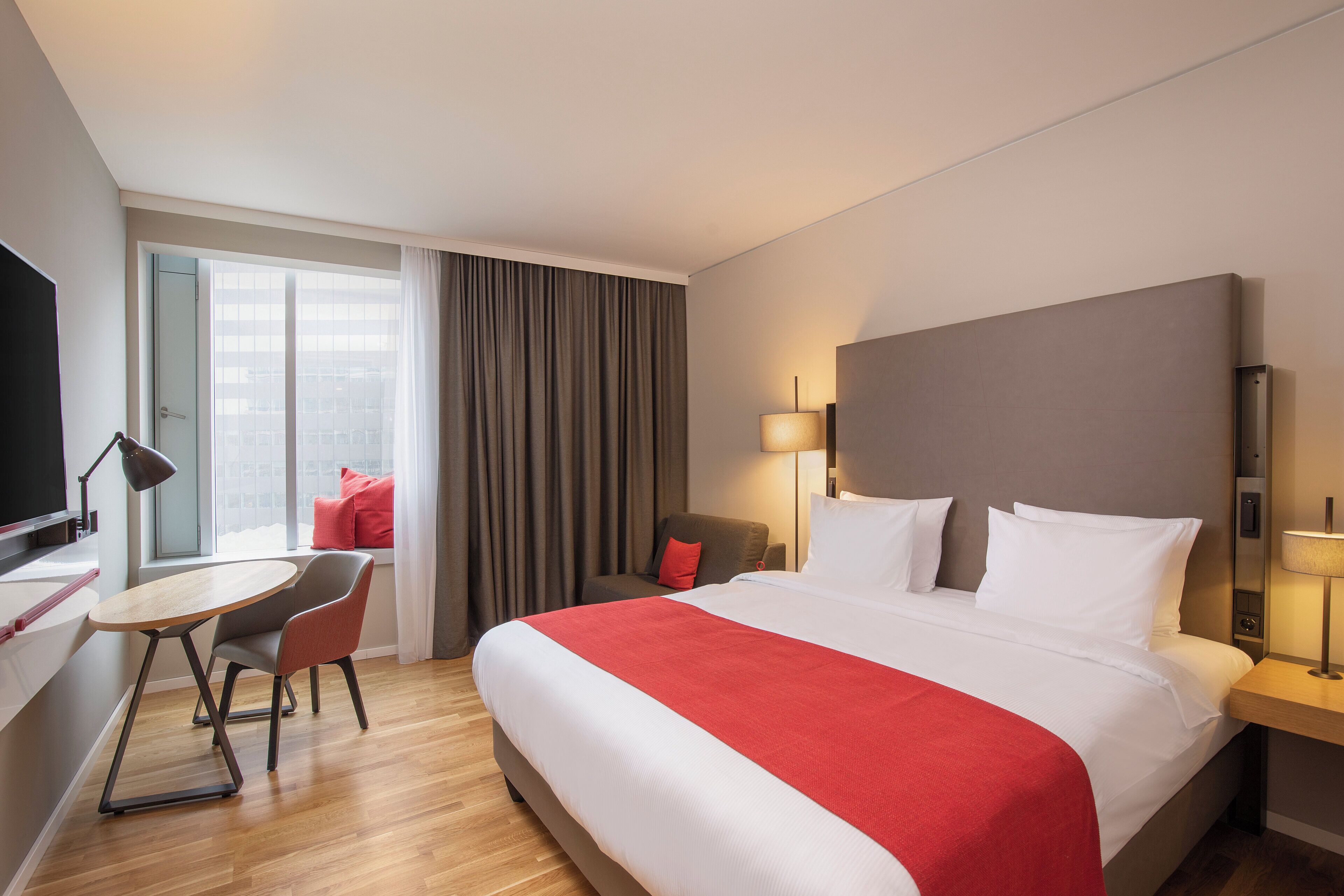 Habitación de hotel con cama, escritorio, silla, televisor y ventana con cortinas.