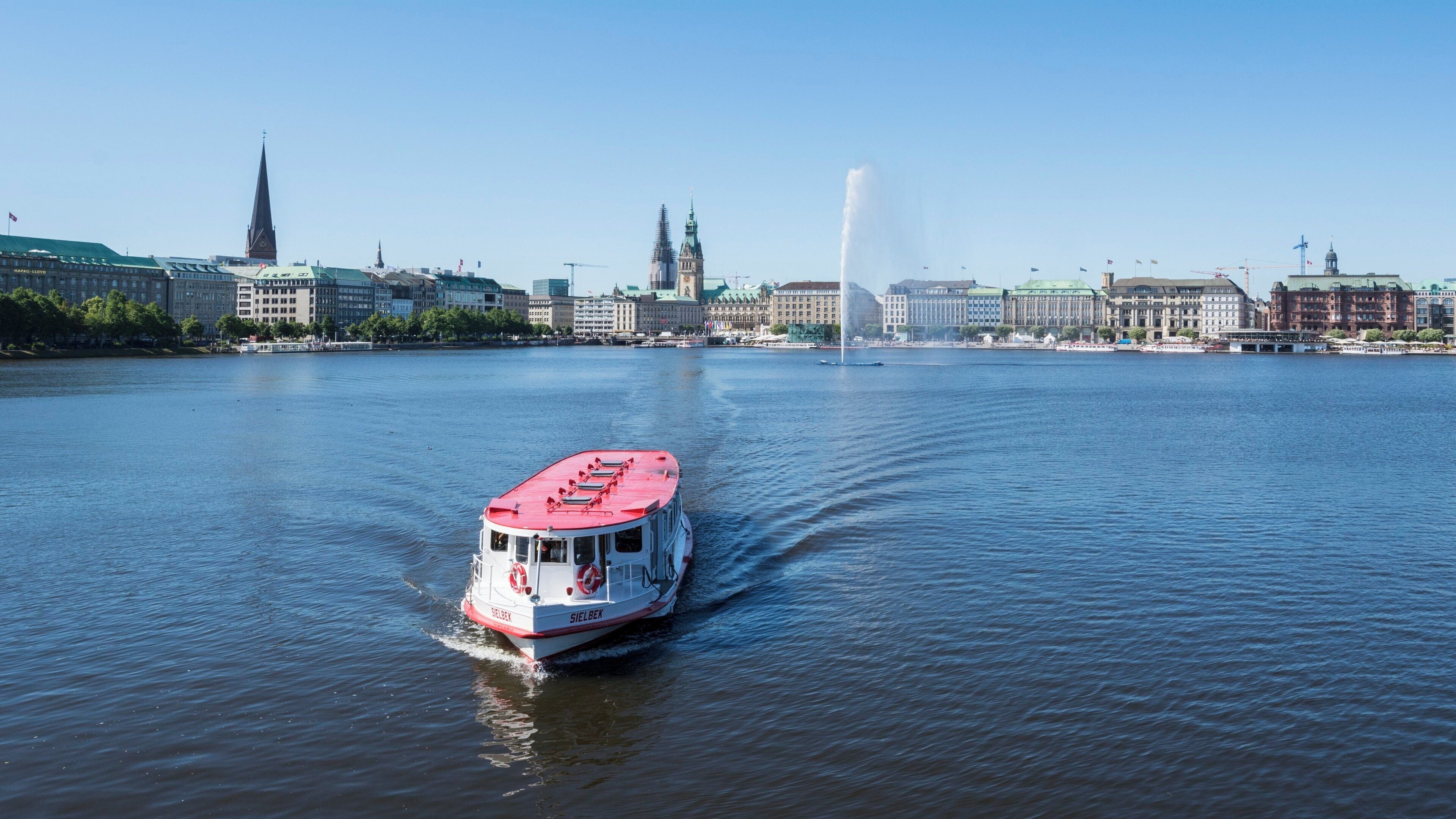 Une rivière sur laquelle navigue un bateau rouge et blanc, se dirigeant vers la silhouette d’une ville, avec ses bâtiments historiques et son imposante tour.