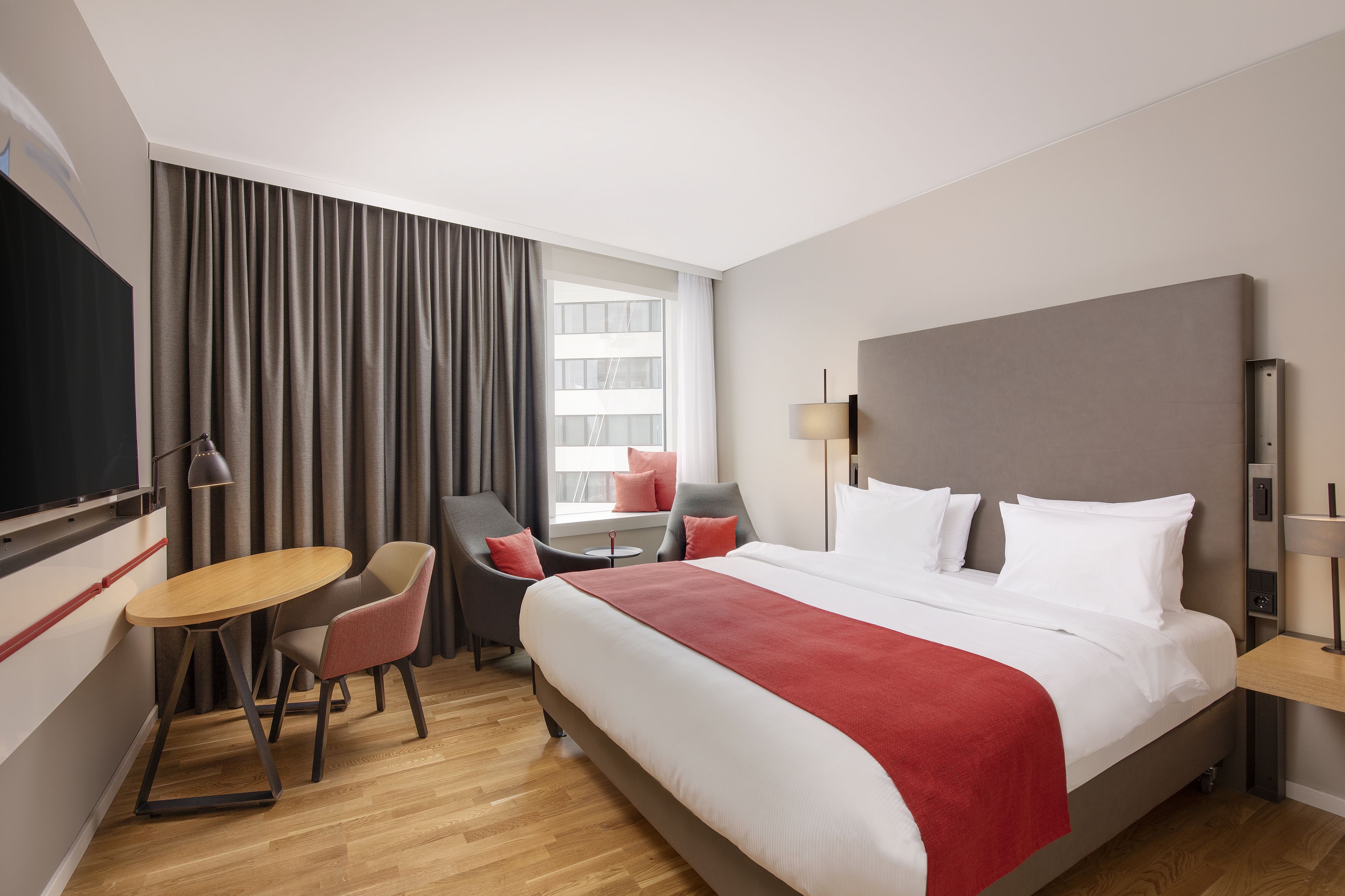 A hotel room with a large bed, a desk with a chair, a TV, and a window with curtains.