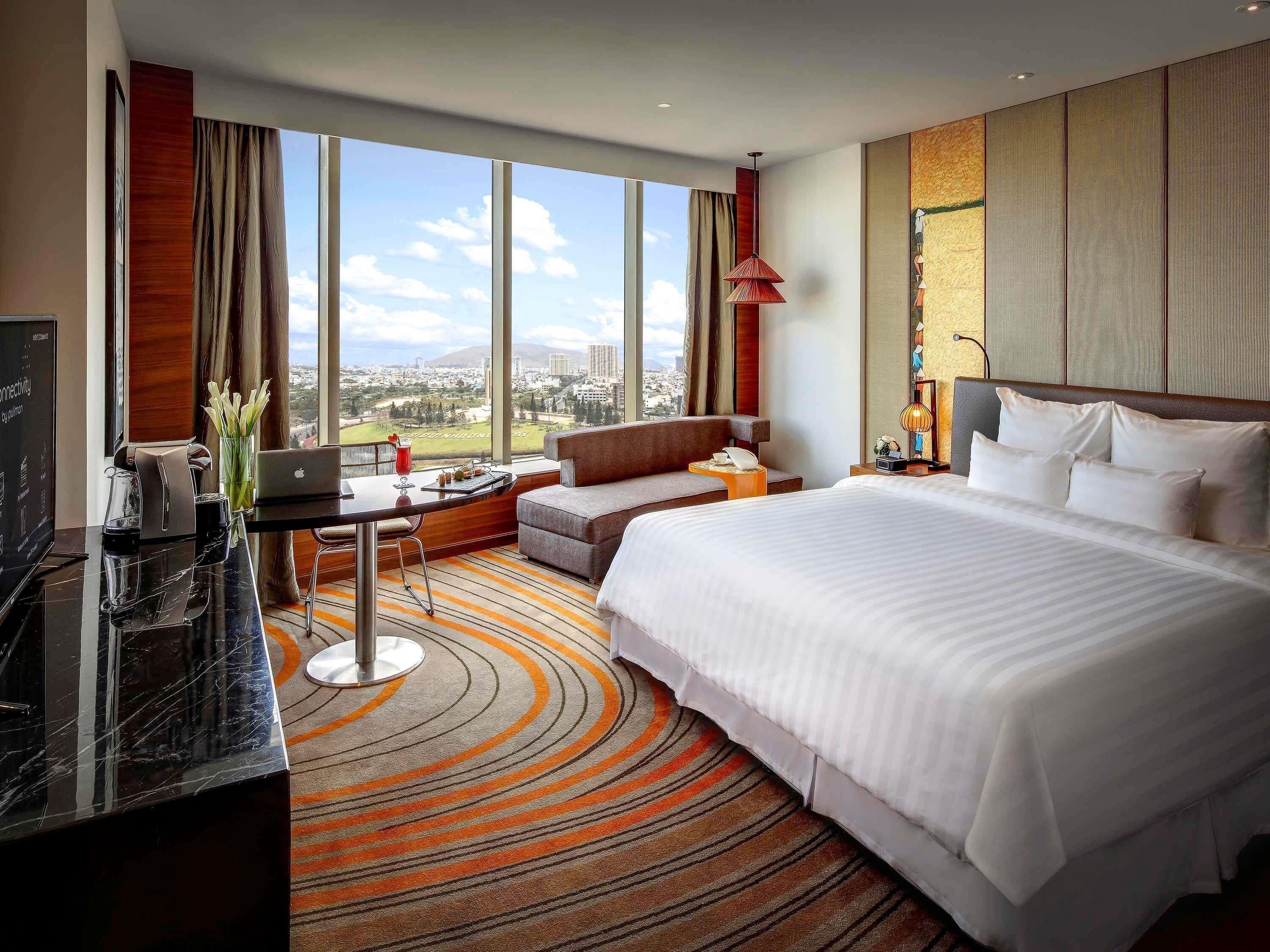 A hotel room with a large bed, a desk with a laptop, a sofa, and a view of the city.