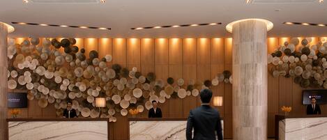A modern hotel lobby with marble reception desks, a large decorative wall, and a tall column.
