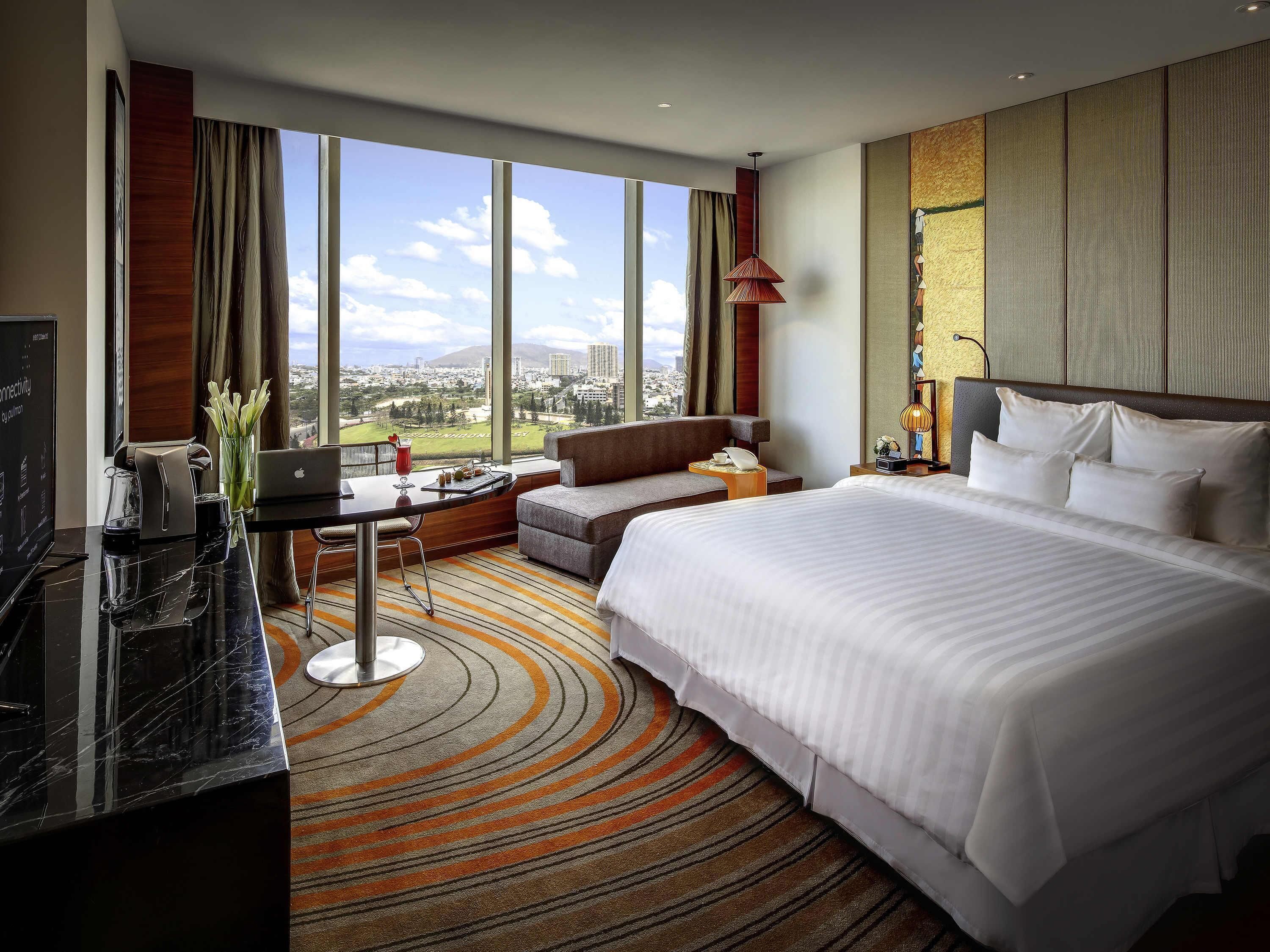 Habitación de hotel con una cama grande, un escritorio con una computadora portátil, vistas a la ciudad y un ventanal amplio.
