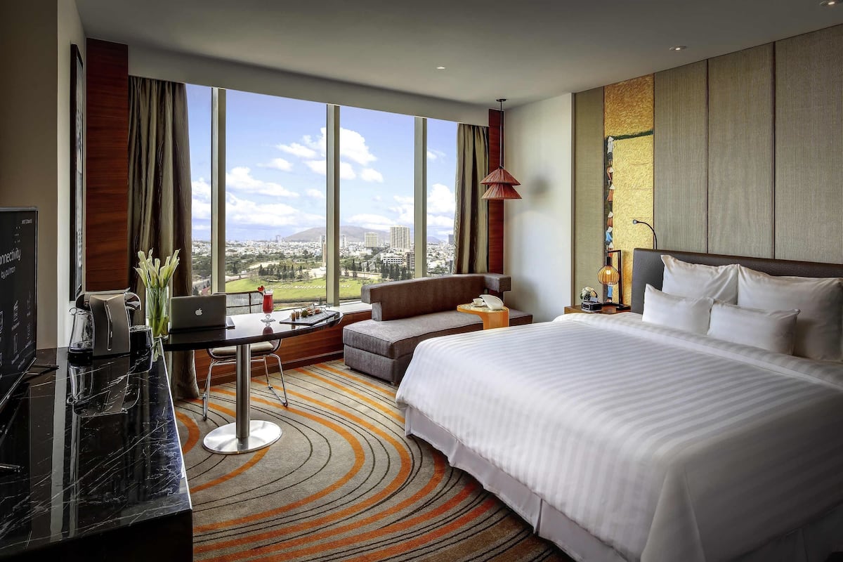 Habitación de hotel con una cama grande, un escritorio con una computadora portátil, vistas a la ciudad y un ventanal amplio.