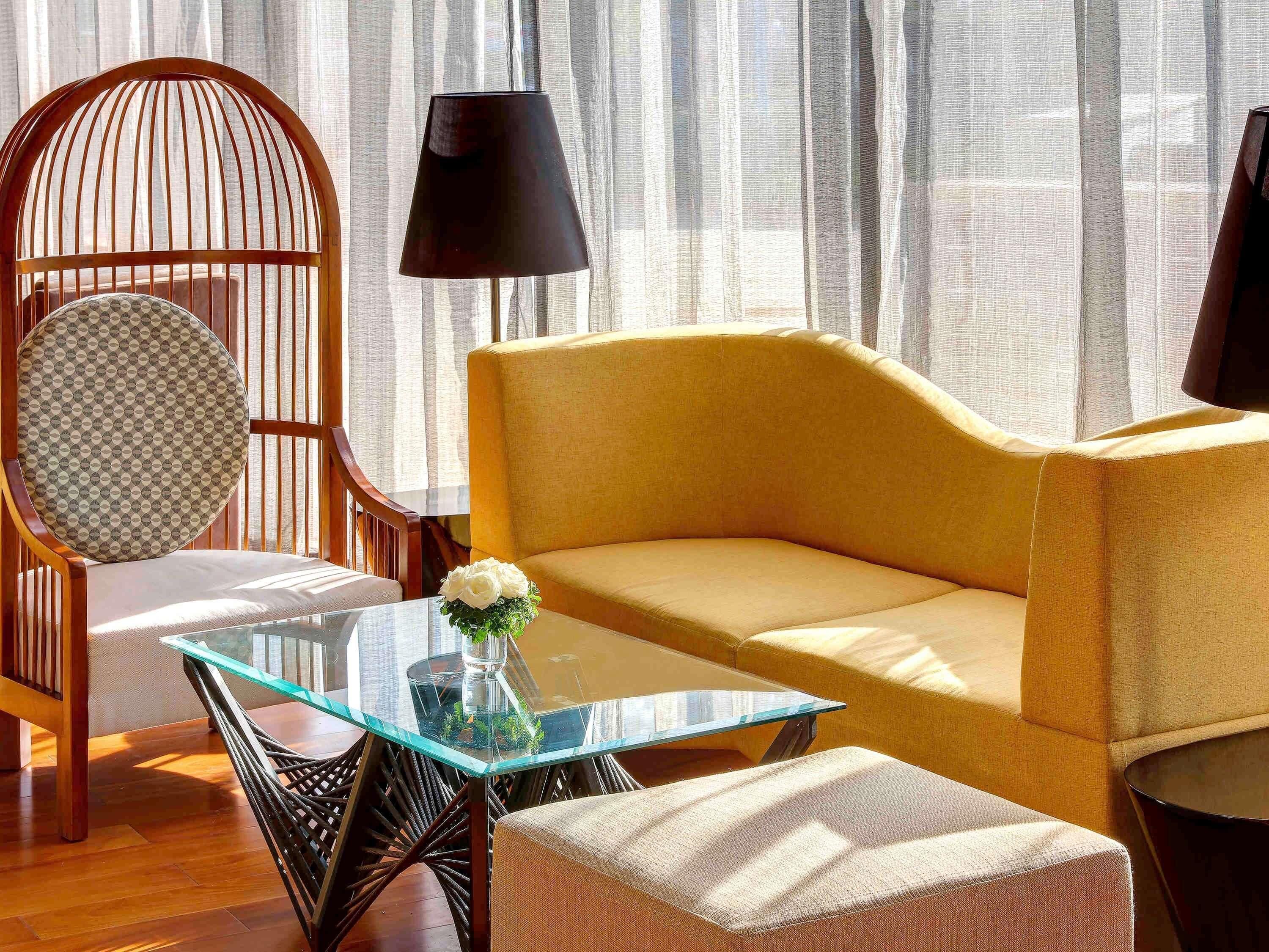 A modern living room with a glass coffee table, a yellow sofa, a wicker chair with a patterned cushion, and a black floor lamp.