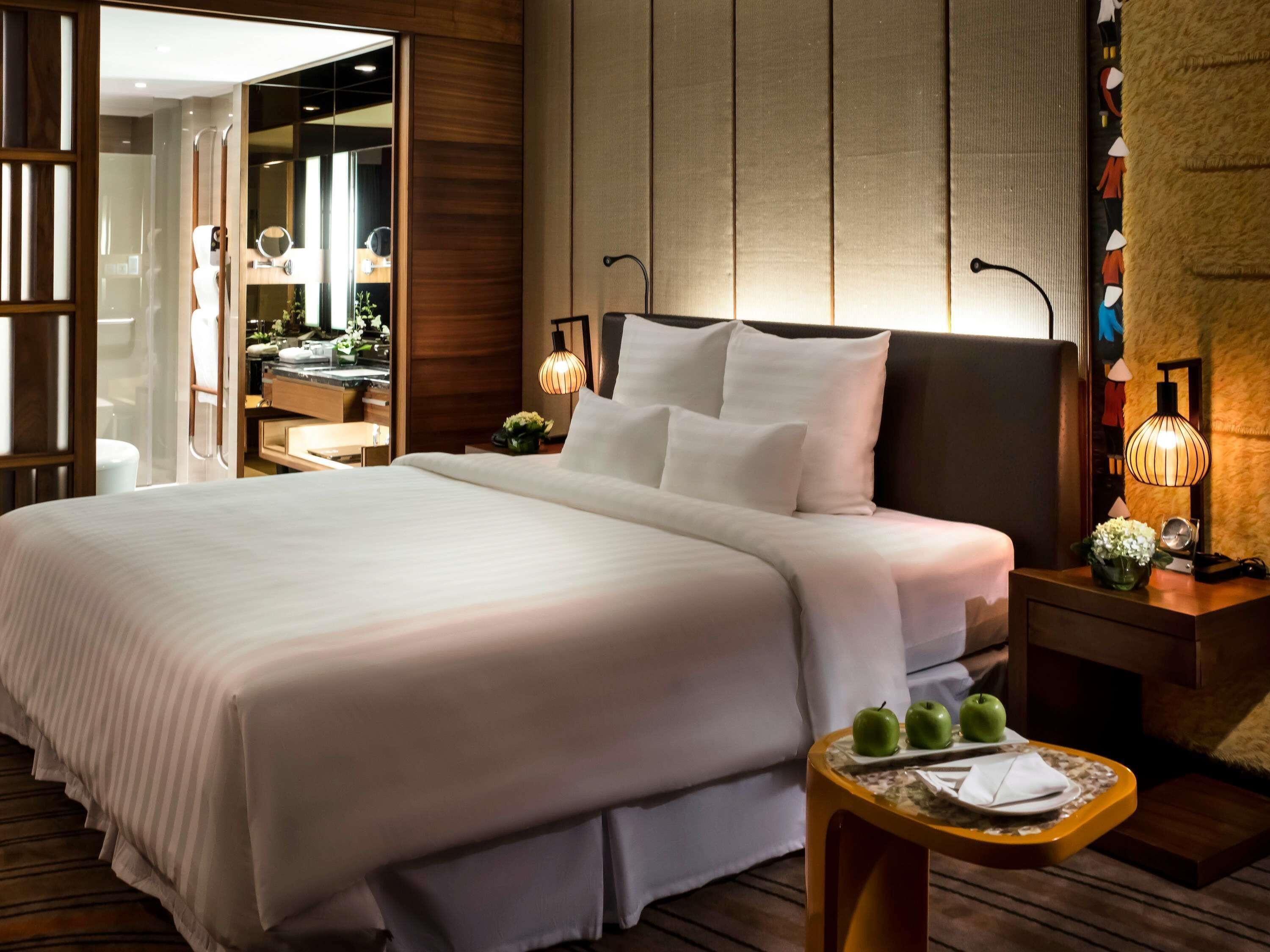 Una habitación de hotel con una cama grande, mesitas de noche, un espejo y una pequeña mesa con manzanas y una servilleta.