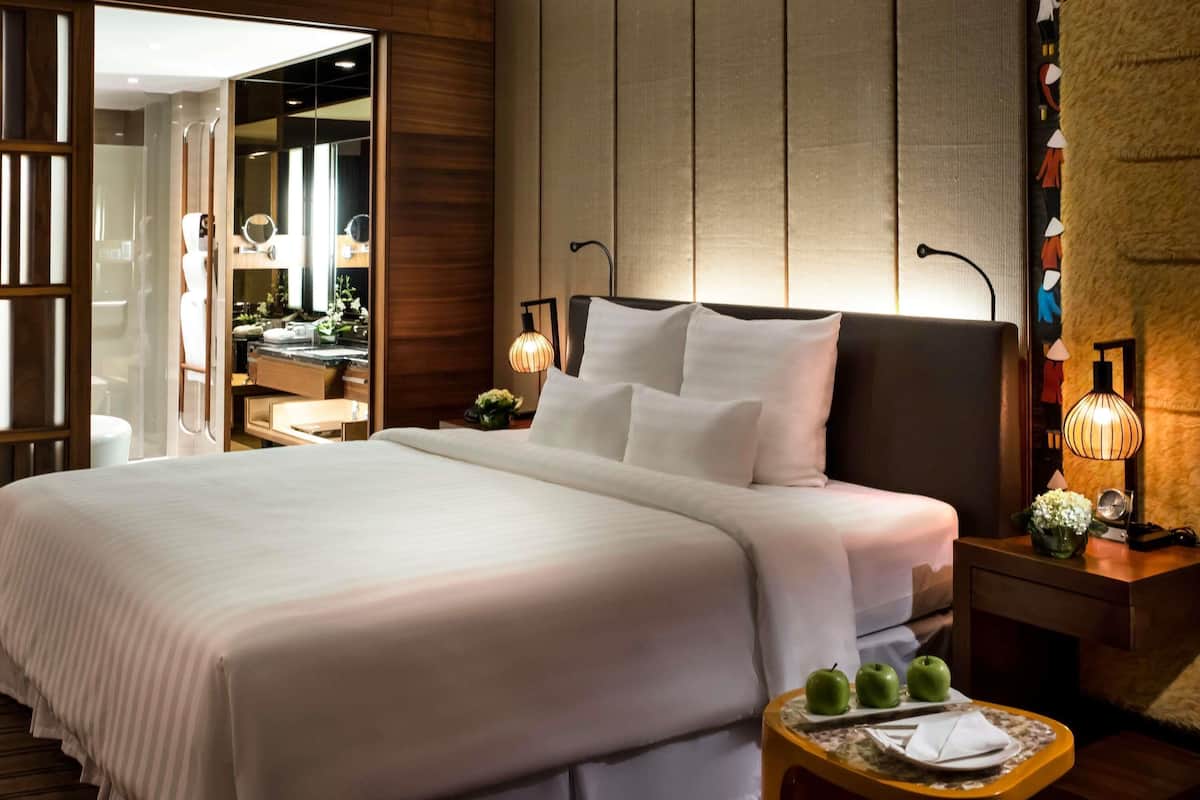Una habitación de hotel con una cama grande, mesitas de noche, un espejo y una pequeña mesa con manzanas y una servilleta.