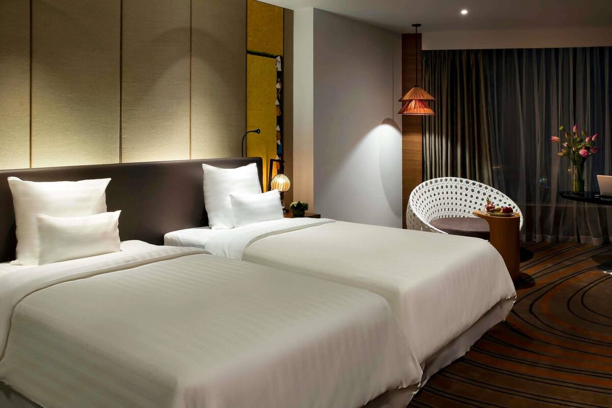 A hotel room with two beds, a white chair, a small table with a vase of flowers, and a view of the city through the window.