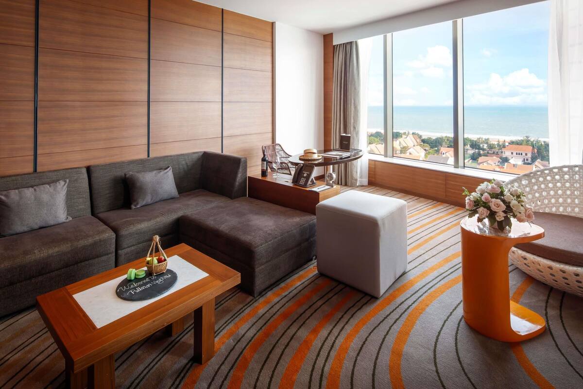 A modern hotel room with a large sofa, a small table with a welcome mat, a round orange side table, and a view of the sea through large windows.