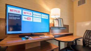 Habitación de hotel con escritorio de madera, televisor de pantalla plana mostrando servicios de streaming, lámpara y una silla.