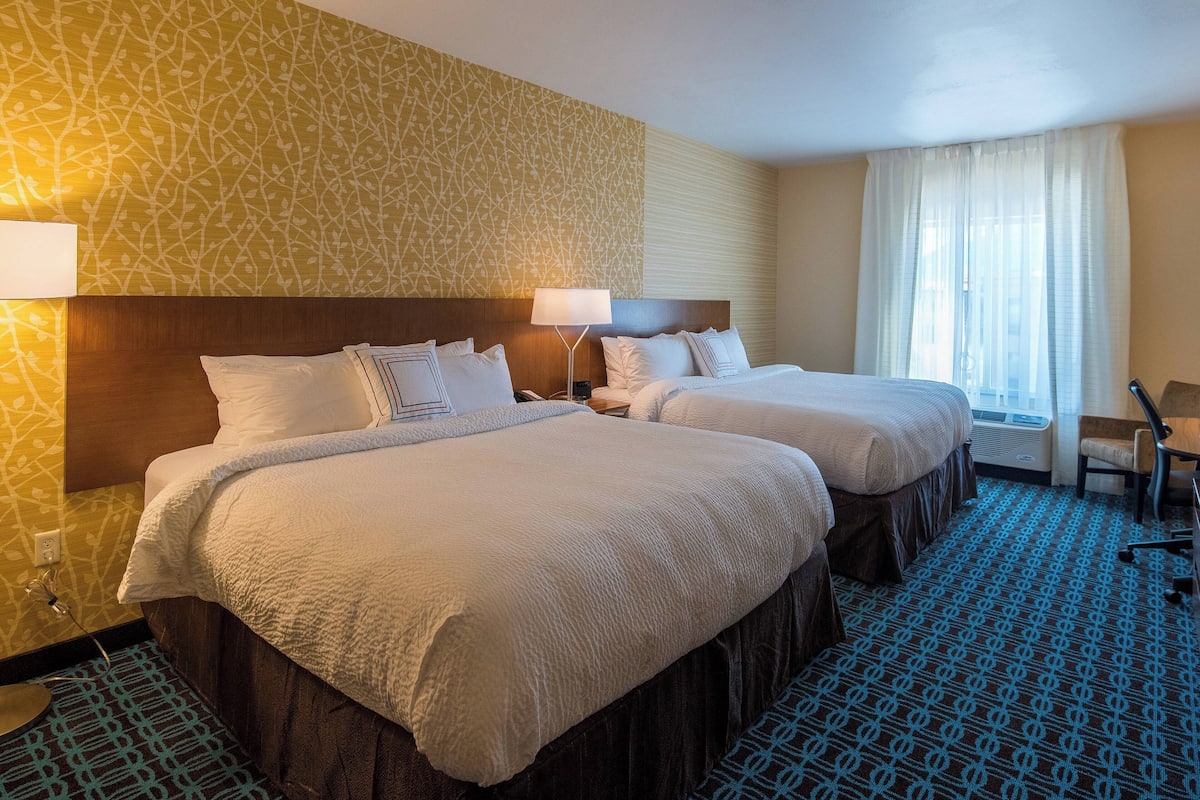 A hotel room with two beds, a wooden headboard, a patterned wall, a large window with curtains, and a patterned carpet.