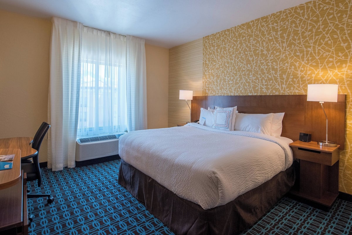 A hotel room with a large bed, two bedside tables with lamps, a desk, a chair, a window with curtains, and a patterned wall.