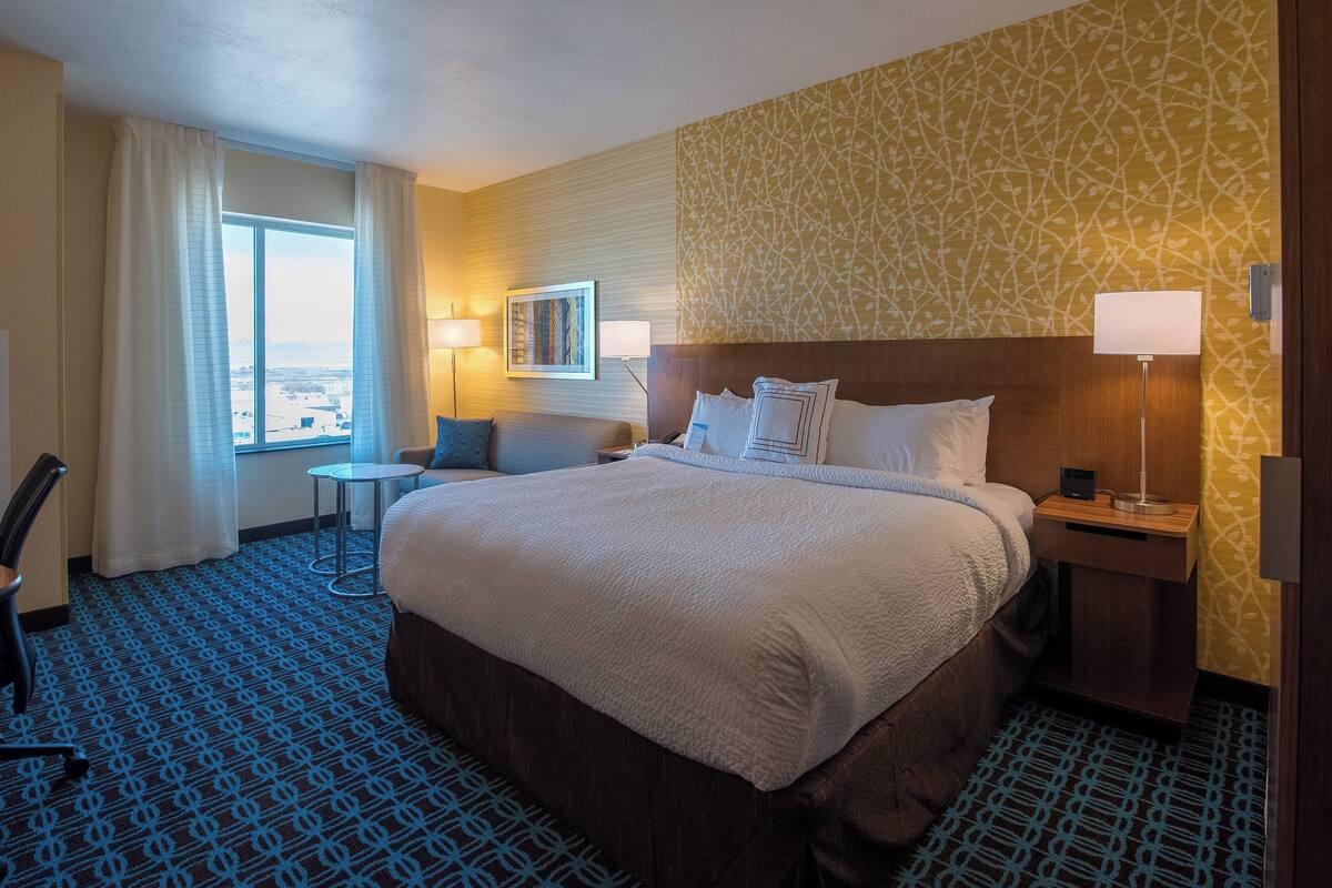 A hotel room with a large bed, a desk, a chair, a window with curtains, and a patterned carpet.