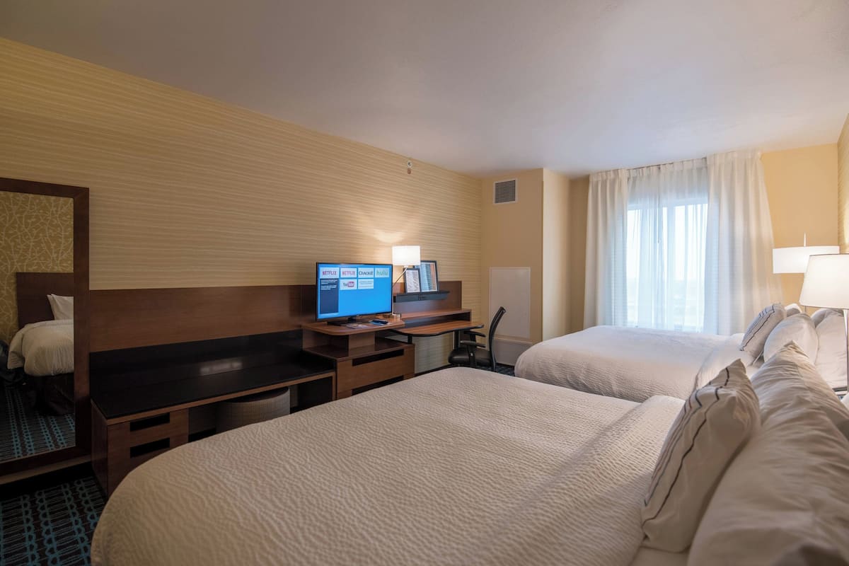 A hotel room with two beds, a desk with a computer, a TV displaying streaming services, and a window with curtains.