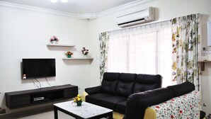 A living room with a black leather sofa, a flat-screen TV mounted on the wall, a coffee table with a vase of flowers, and a window with floral curtains. - Garden Vacation Suite (Bentong)