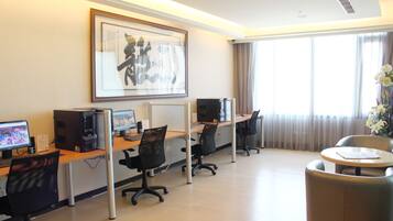 A hotel room with a desk, computer, and chairs. There is a large framed artwork on the wall and a window with curtains.