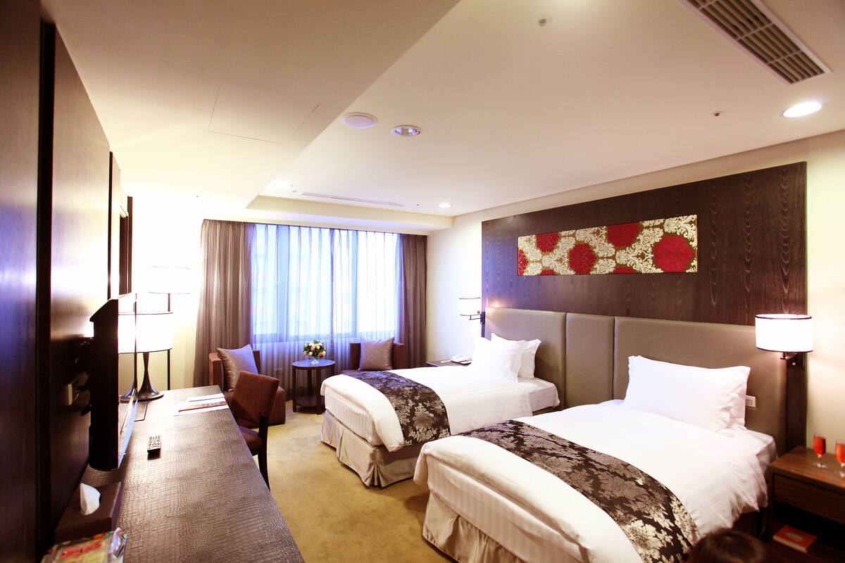 A hotel room with two beds, a desk, a chair, a small table with a lamp, and a window with curtains.
