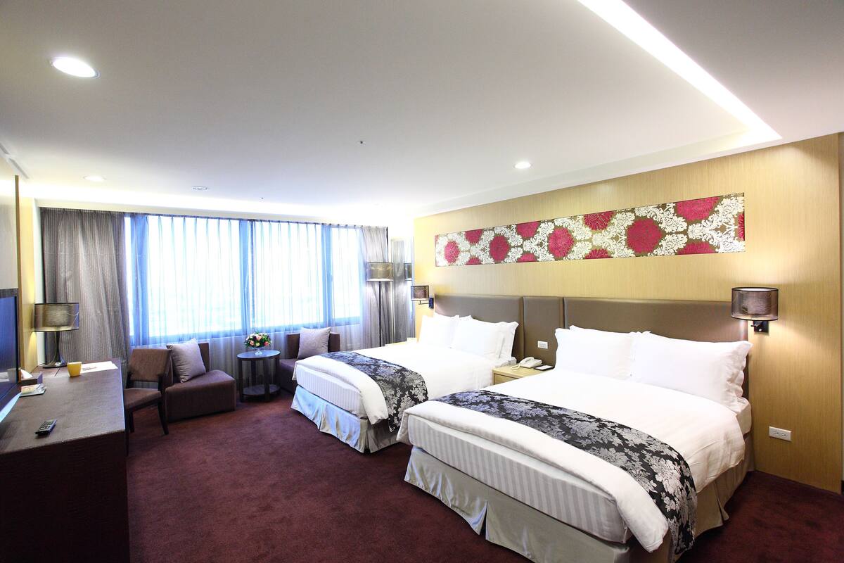 A hotel room with two beds, a desk, a chair, a small table, and a window with curtains.