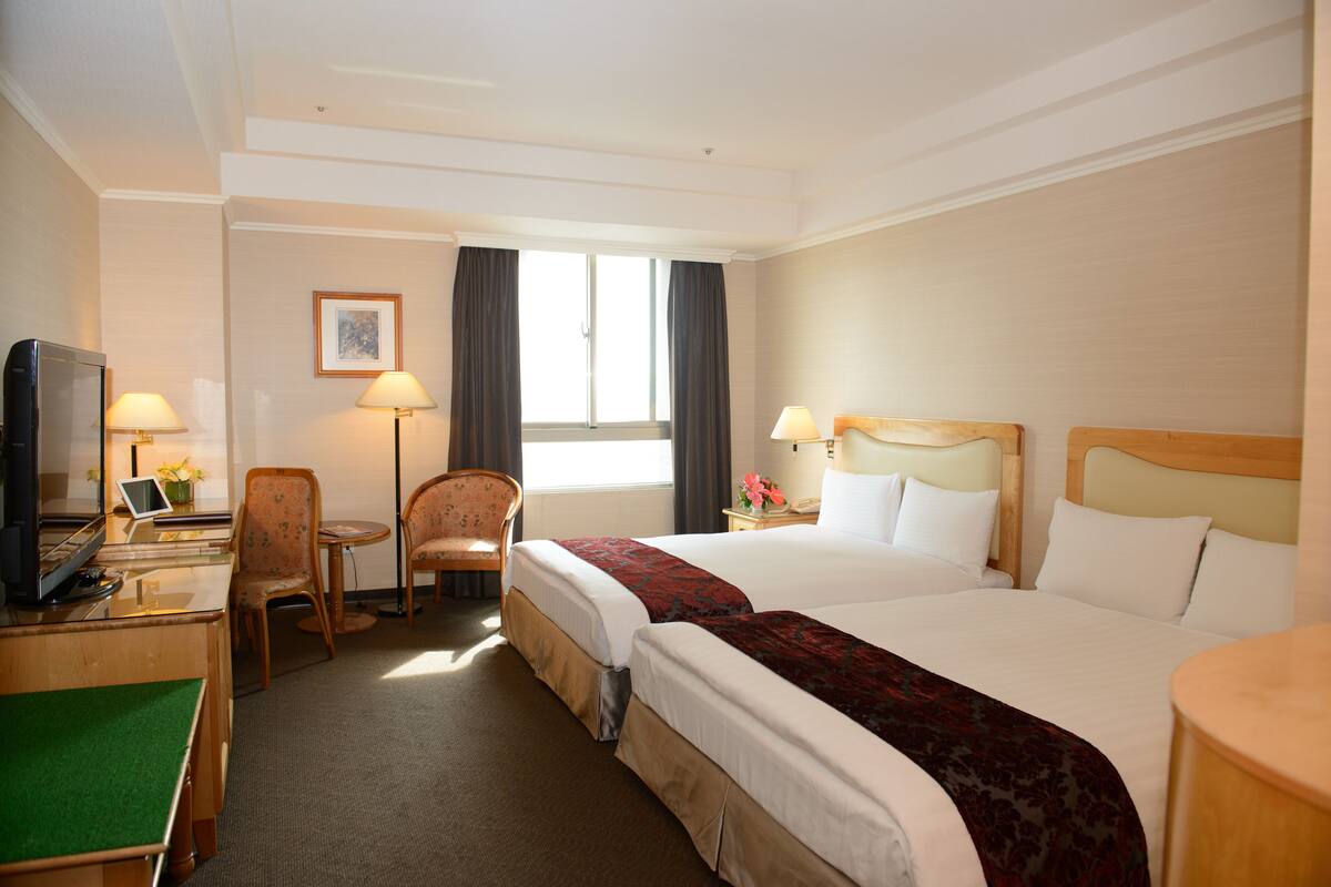 A hotel room with two beds, a desk with a computer, a chair, and a window with curtains.
