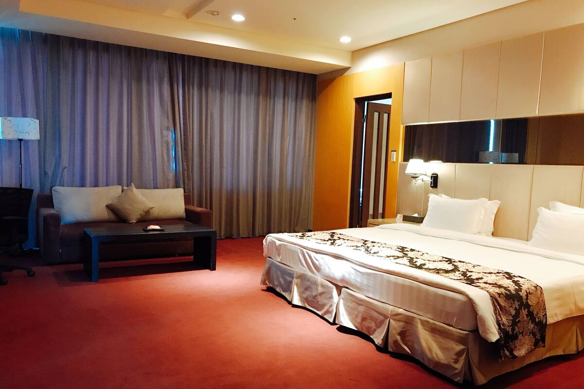 A hotel room with a bed, a sofa, a desk, and a large window with curtains.
