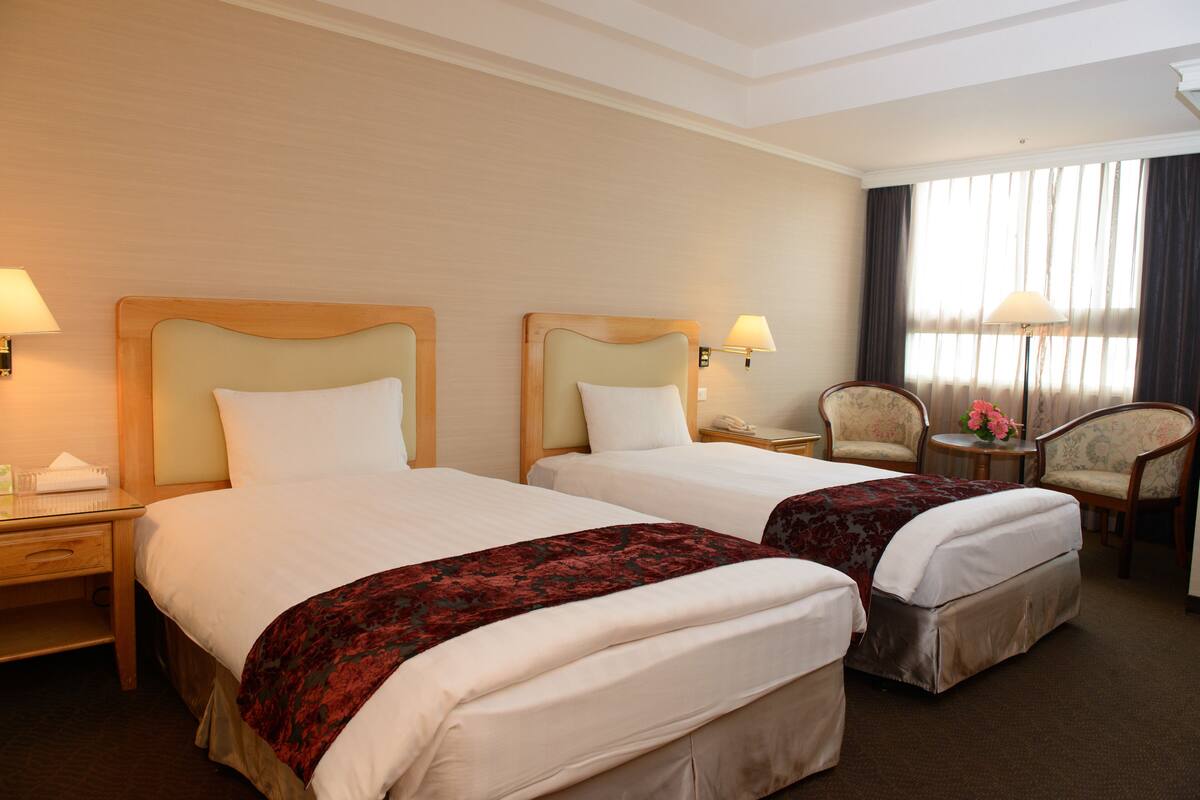A hotel room with two beds, a desk, a chair, and a window with curtains.