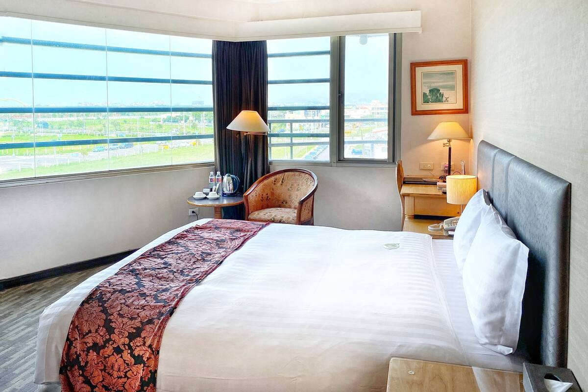 A hotel room with a large bed, a chair, a desk, a lamp, and a view of the city through large windows.