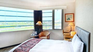 A hotel room with a large bed, a chair, a desk, a lamp, and a view of the city through large windows.
