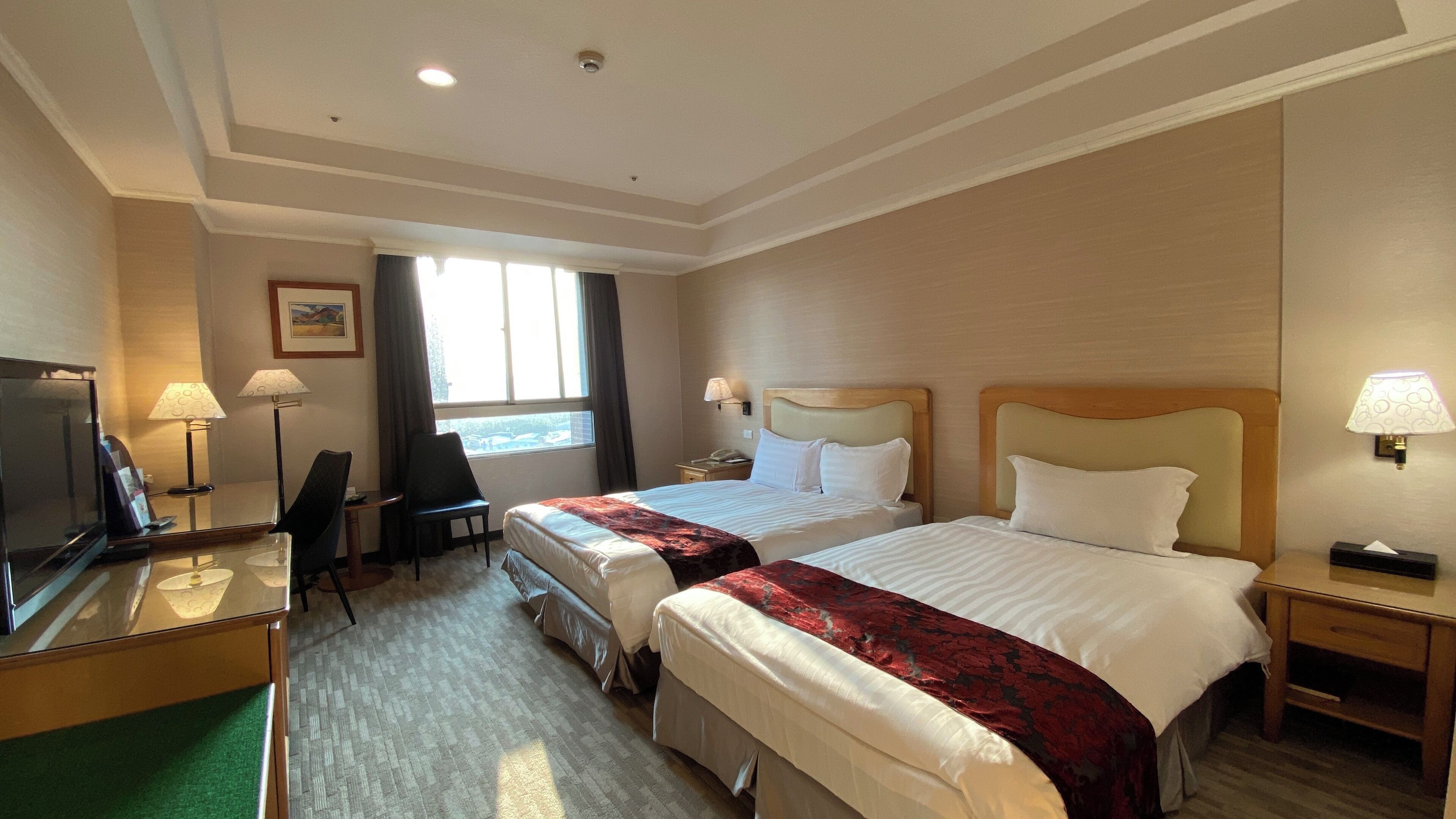 A hotel room with two beds, a desk with a TV, a chair, a window with curtains, and a lamp on the nightstand.