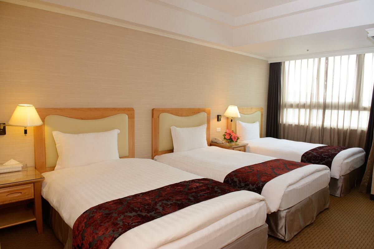 A hotel room with two beds, each with a wooden headboard, a bedside table with a lamp, and a window with curtains.