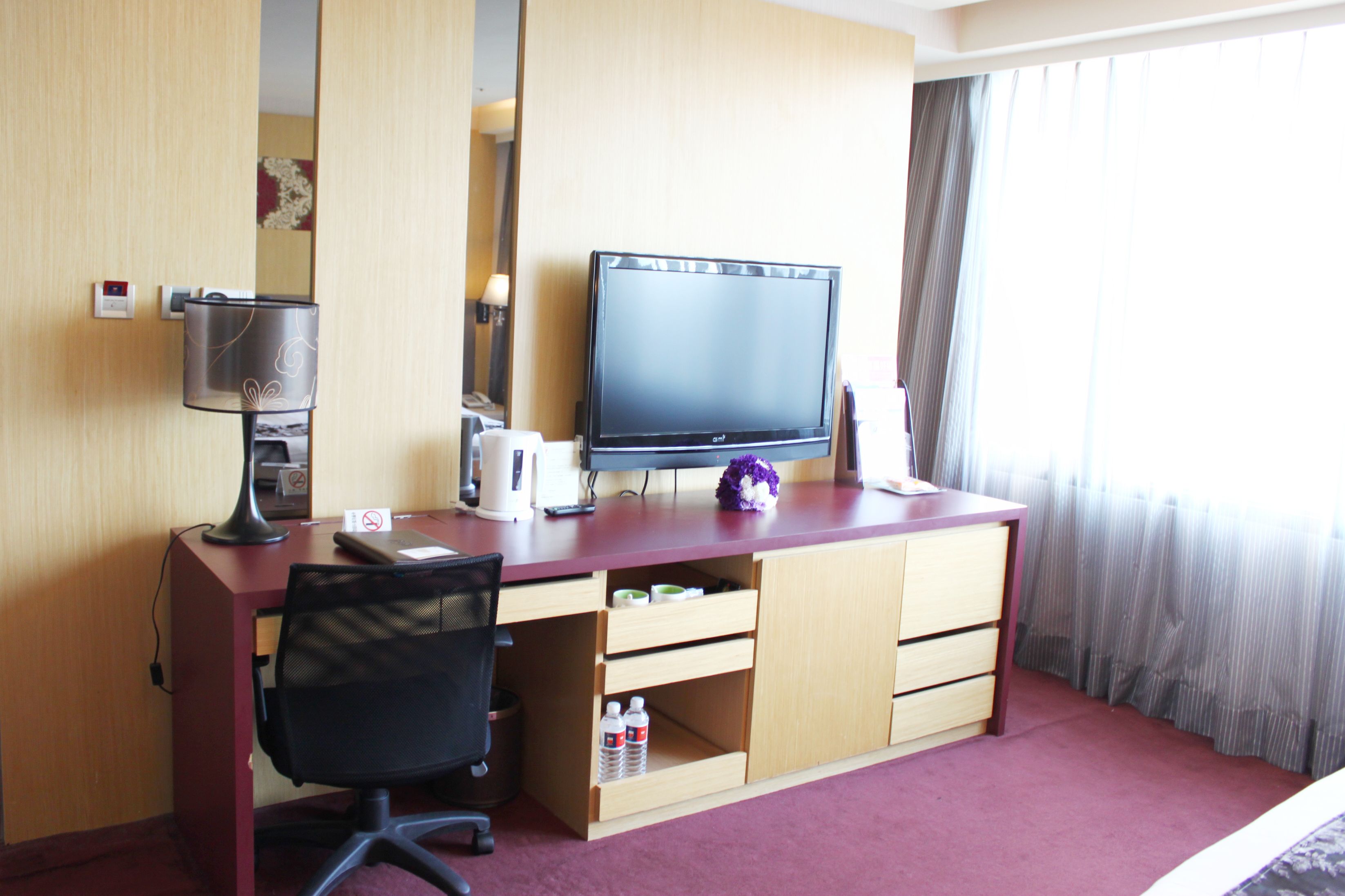 A hotel room with a wooden desk, a black office chair, a flat-screen TV, a lamp, a coffee maker, and a bottle of water.