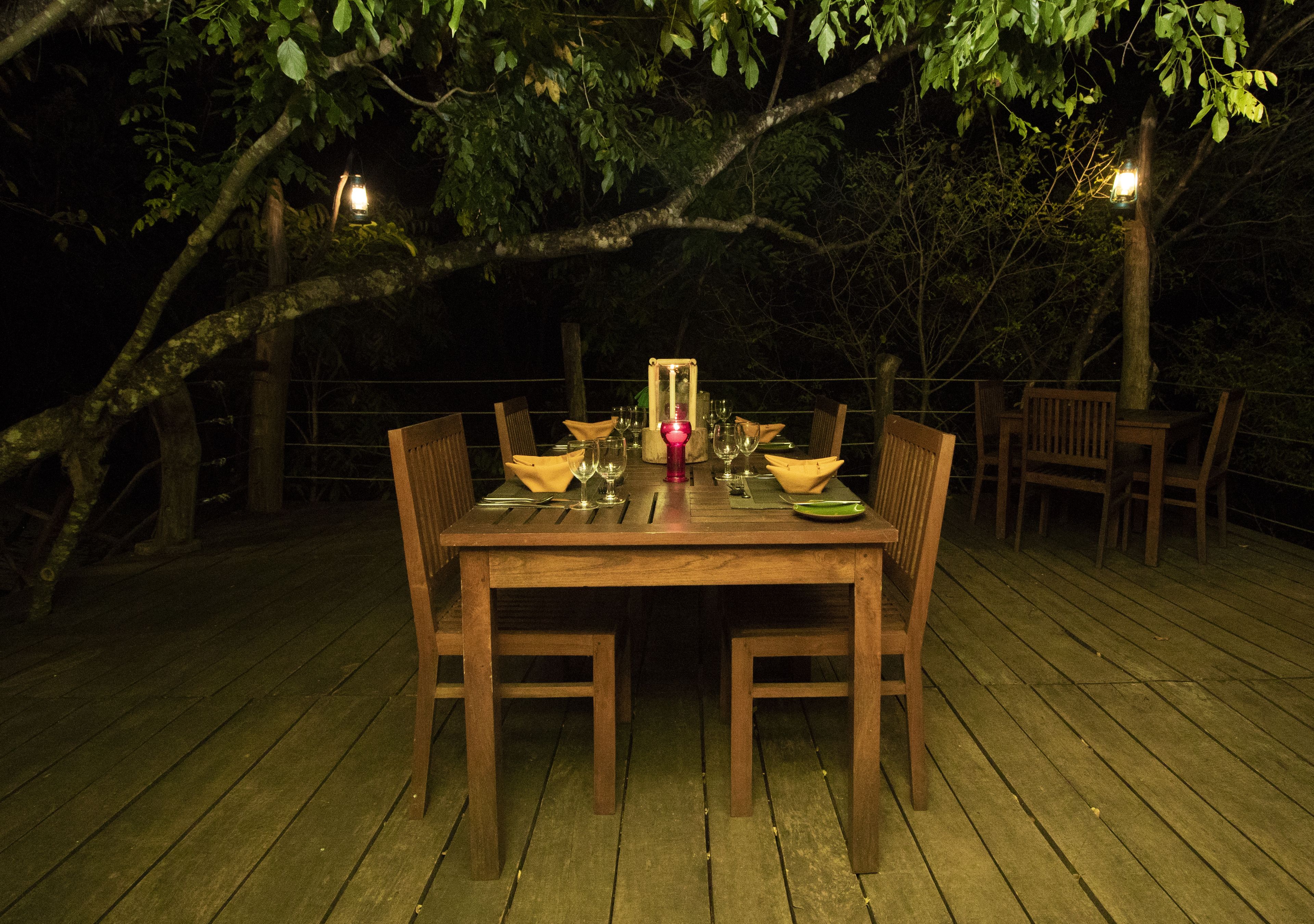 Una mesa de comedor de madera al aire libre con sillas, rodeada de árboles y una terraza de madera.