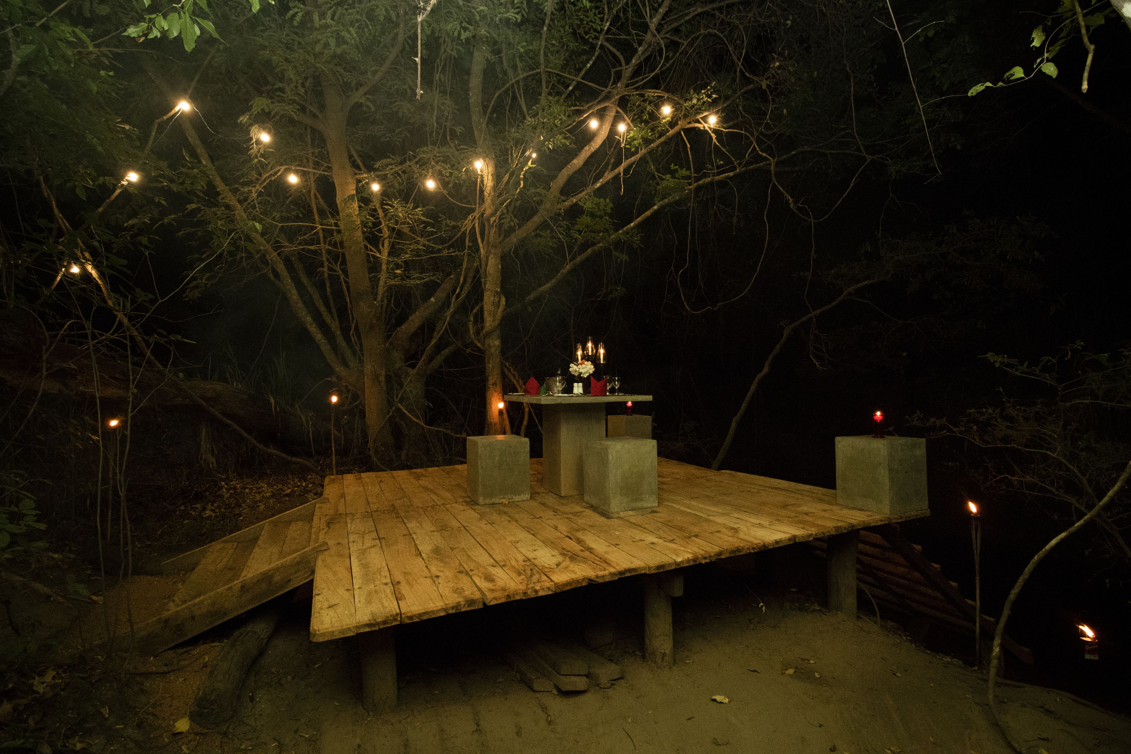 A wooden platform with candles and a small table, surrounded by trees and string lights.