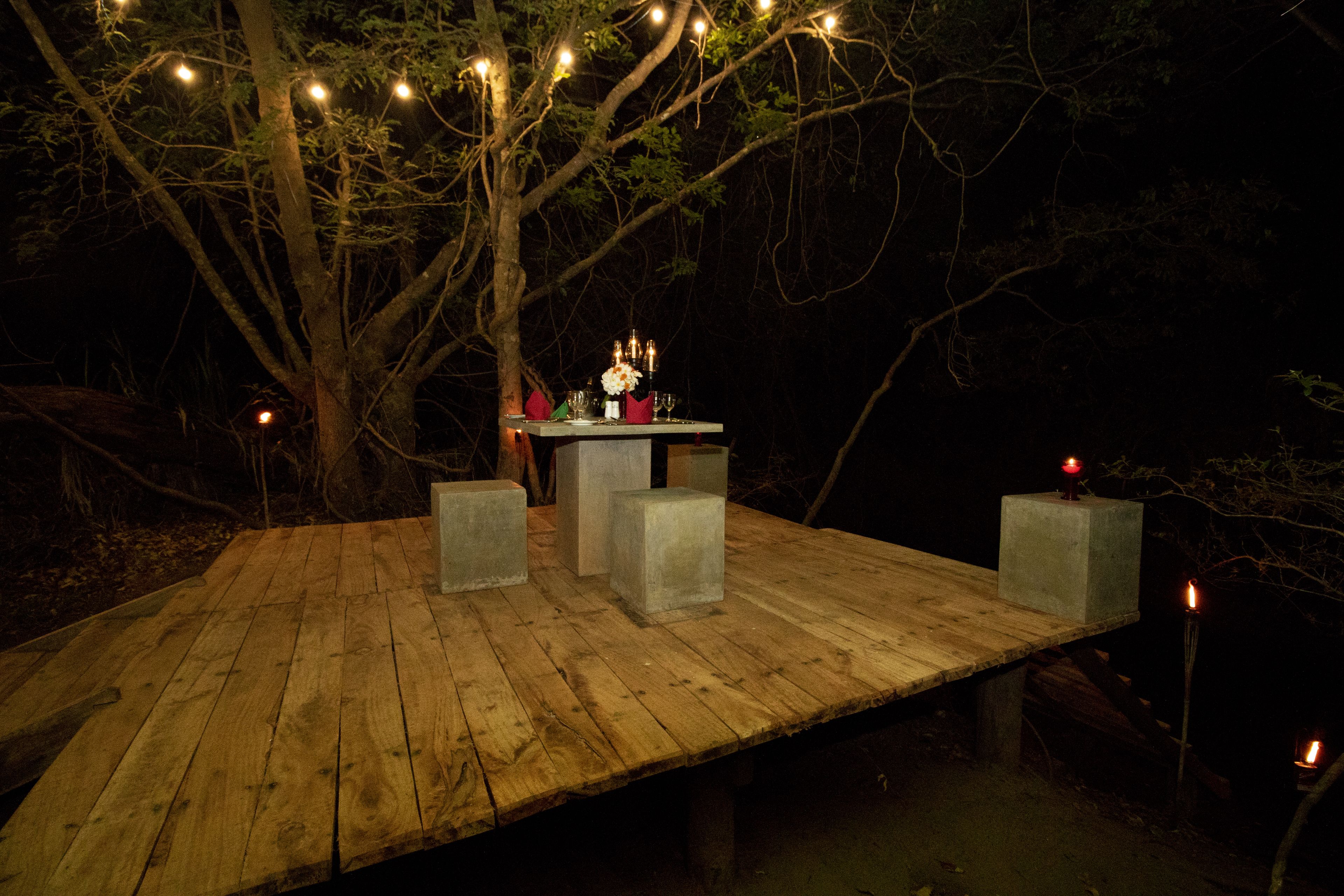 A wooden deck with a table set for a meal, surrounded by trees and string lights.