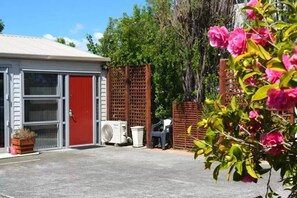 A building with a red door, a white wall, and a window. There is a wooden fence and a tree with pink flowers in the foreground. - McWilliam Apartments (Christchurch)